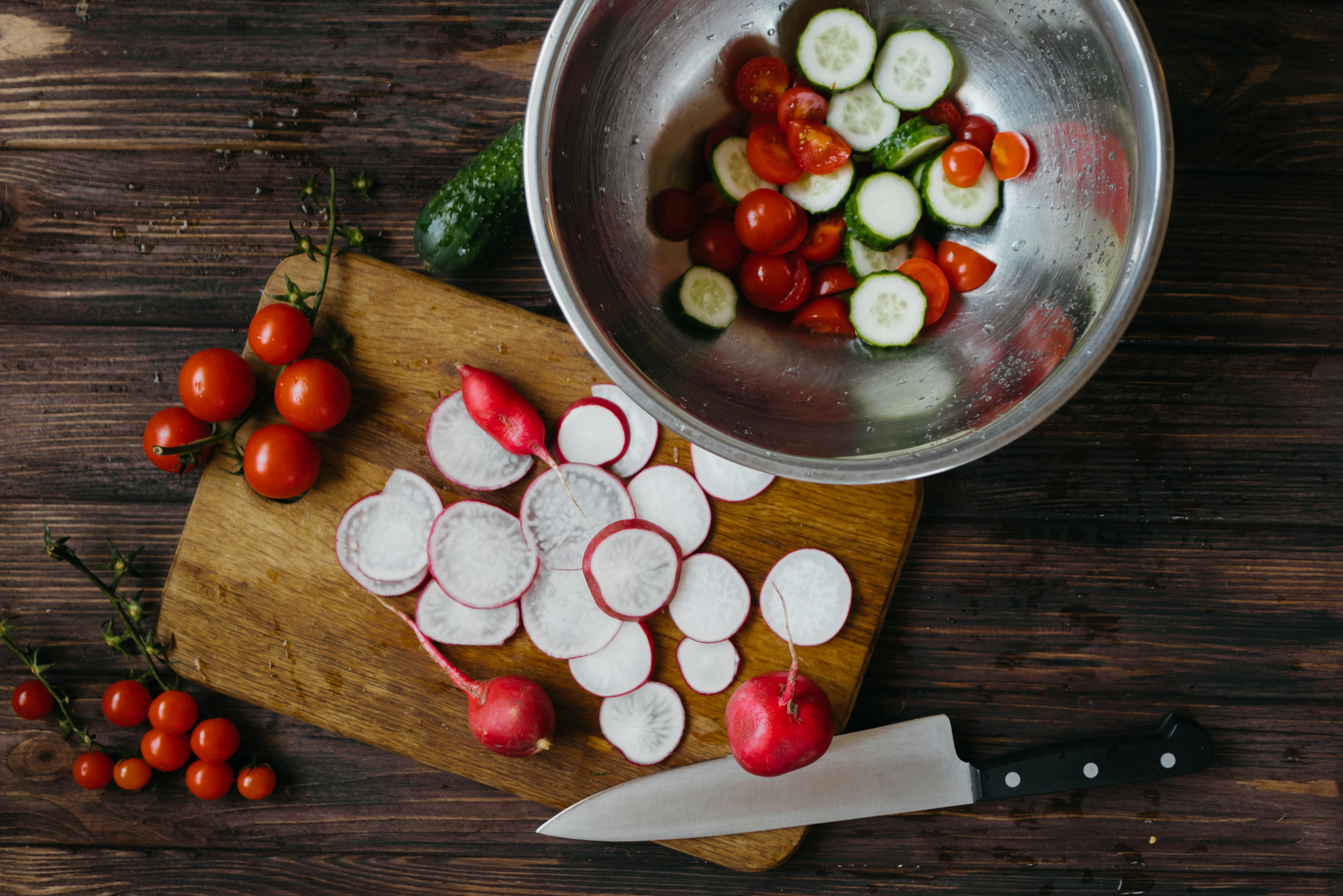 Radishes on a cutting board next to a bowl with cucumbers and tomatoes